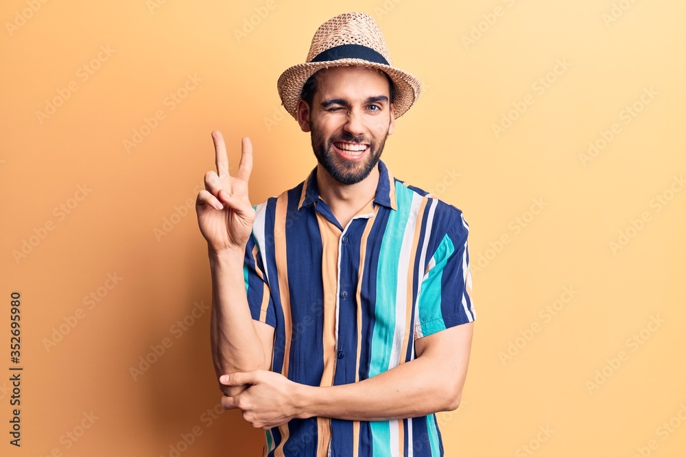Young handsome man with beard wearing summer hat and shirt smiling with happy face winking at the camera doing victory sign. number two.