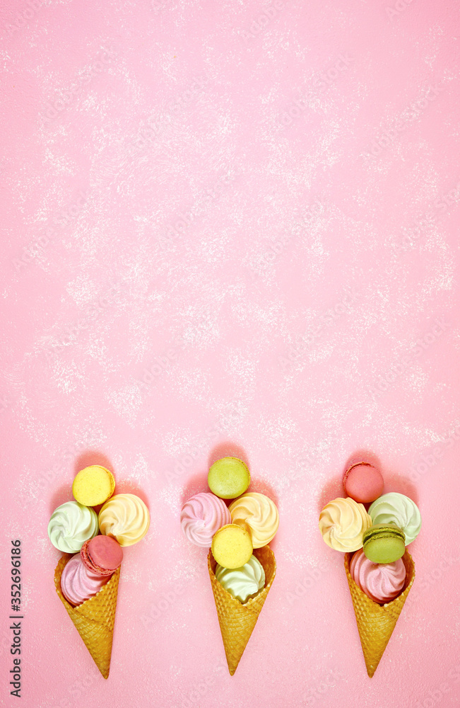 Summertime decorated border of ice cream cones filled with macarons and meringues on pink textured background, negative copy space. Stylish flat lay, top view minimalism creative layout. Vertical.