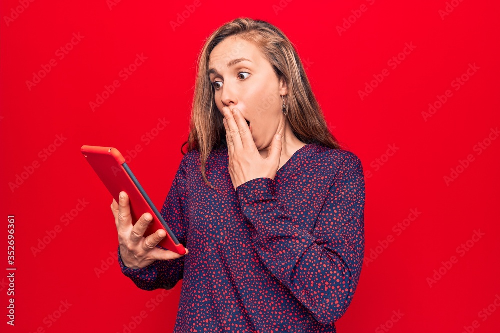 Naklejka premium Young beautiful blonde woman using tablet over isolated red background covering mouth with hand, shocked and afraid for mistake. surprised expression