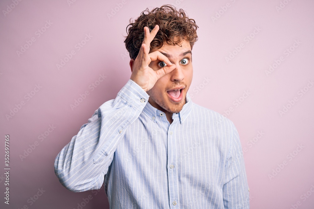Young blond handsome man with curly hair wearing striped shirt over white background doing ok gesture with hand smiling, eye looking through fingers with happy face. Young blond handsome man with curly hair wearing striped shirt over white background doing ok gesture with hand smiling, eye looking through fingers with happy face.