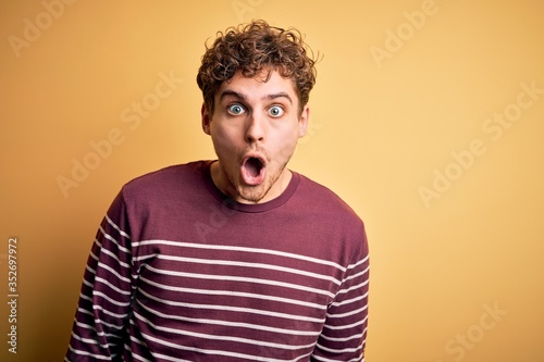 Young blond handsome man with curly hair wearing casual striped sweater afraid and shocked with surprise expression, fear and excited face.