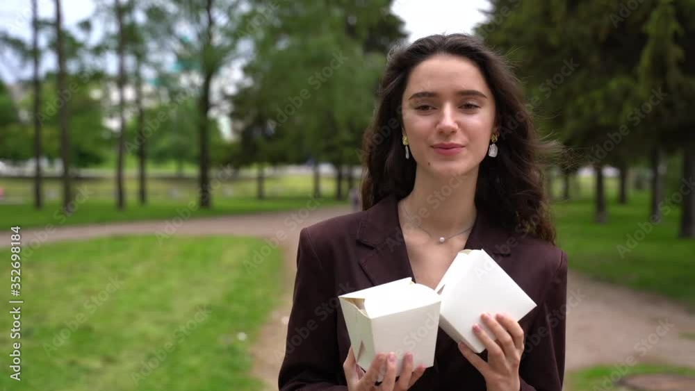 Girl wakls and smiles in park and holds wok in box udon noodles with tempuru, shrimps, in hands. 4k video Udon noodles in white box delivery. 2 Wok box udon advertise 1+1. Girl wakls and smiles in park and holds wok in box udon noodles with tempuru, shrimps, in hands. 4k video Udon noodles in white box delivery. 2 Wok box udon advertise 1+1.