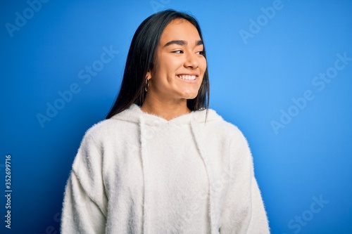 Young beautiful asian sportswoman wearing sweatshirt standing over isolated blue background looking away to side with smile on face, natural expression. Laughing confident.