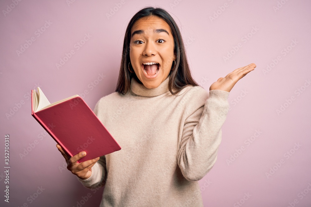 Young asian smart woman holding book diary standing over isolated pink background very happy and excited, winner expression celebrating victory screaming with big smile and raised hands Young asian smart woman holding book diary standing over isolated pink background very happy and excited, winner expression celebrating victory screaming with big smile and raised hands