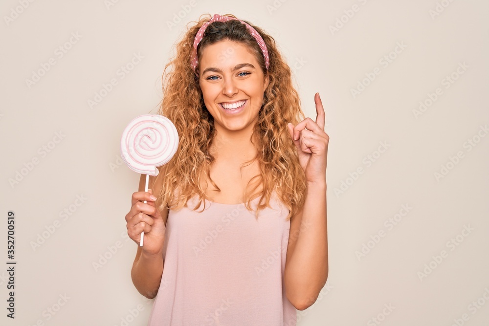 Young beautiful blonde woman with blue eyes eating sweet candy lollipop over pink background surprised with an idea or question pointing finger with happy face, number one Young beautiful blonde woman with blue eyes eating sweet candy lollipop over pink background surprised with an idea or question pointing finger with happy face, number one