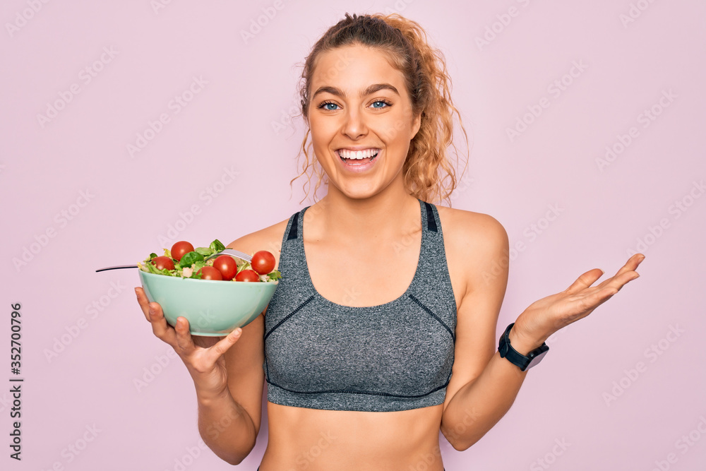 Young beautiful blonde sporty woman with blue eyes holding bowl with healthy salad very happy and excited, winner expression celebrating victory screaming with big smile and raised hands Young beautiful blonde sporty woman with blue eyes holding bowl with healthy salad very happy and excited, winner expression celebrating victory screaming with big smile and raised hands