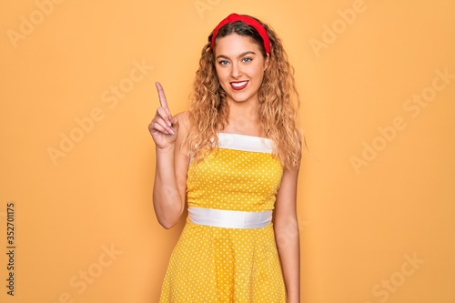 Beautiful blonde pin-up woman with blue eyes wearing diadem standing over yellow background pointing finger up with successful idea. Exited and happy. Number one.