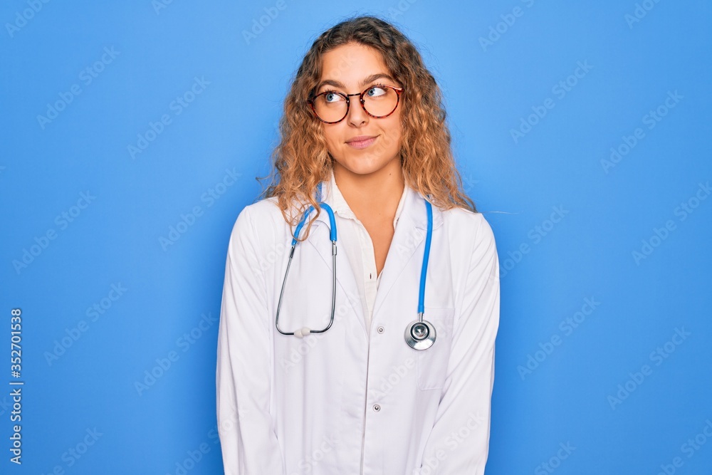 Young beautiful blonde doctor woman with blue eyes wearing coat and stethoscope smiling looking to the side and staring away thinking. Young beautiful blonde doctor woman with blue eyes wearing coat and stethoscope smiling looking to the side and staring away thinking.