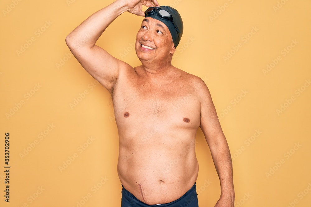 Middle age senior grey-haired swimmer man wearing swimsuit, cap and goggles smiling confident touching hair with hand up gesture, posing attractive and fashionable