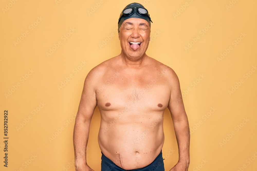 Middle age senior grey-haired swimmer man wearing swimsuit, cap and goggles sticking tongue out happy with funny expression. Emotion concept.