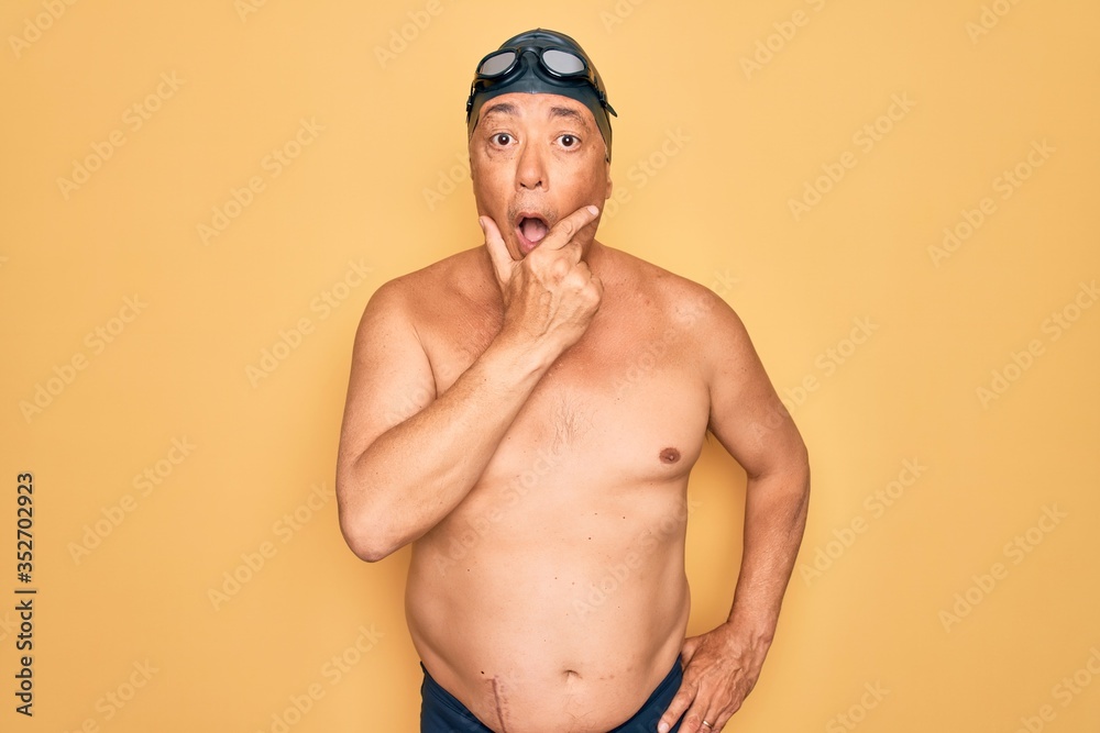 Middle age senior grey-haired swimmer man wearing swimsuit, cap and goggles Looking fascinated with disbelief, surprise and amazed expression with hands on chin