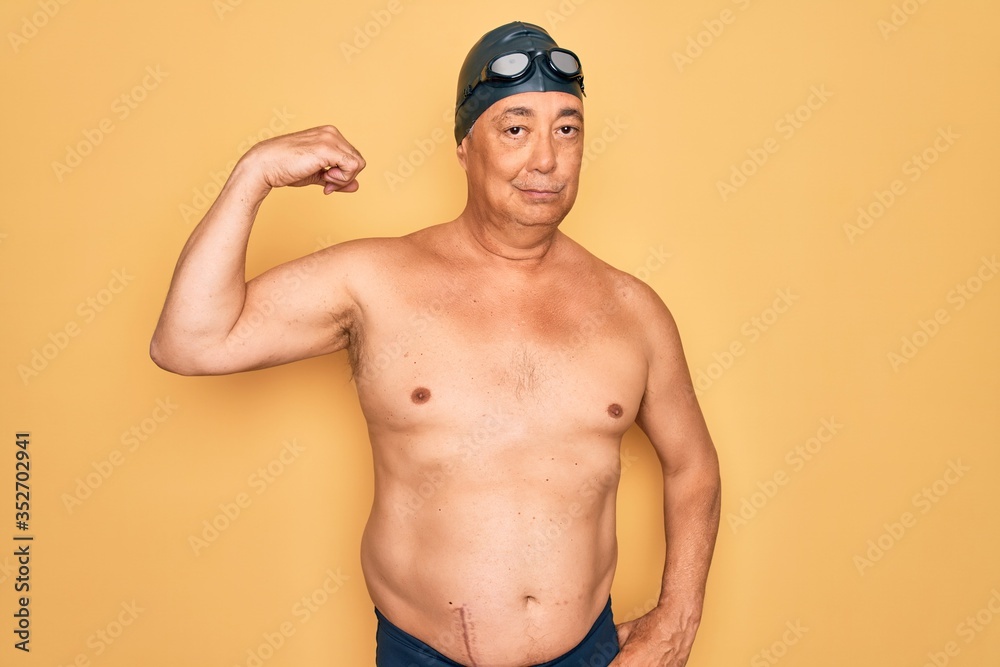 Middle age senior grey-haired swimmer man wearing swimsuit, cap and goggles Strong person showing arm muscle, confident and proud of power