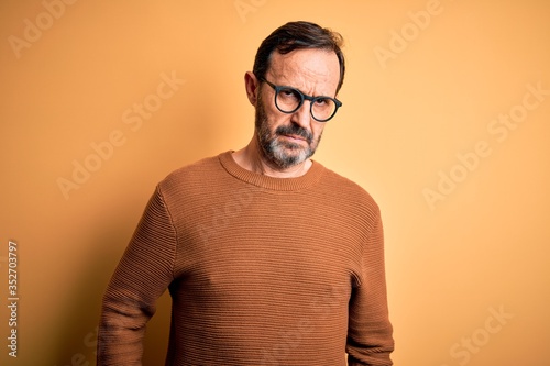 Middle age hoary man wearing brown sweater and glasses over isolated yellow background skeptic and nervous, frowning upset because of problem. Negative person.