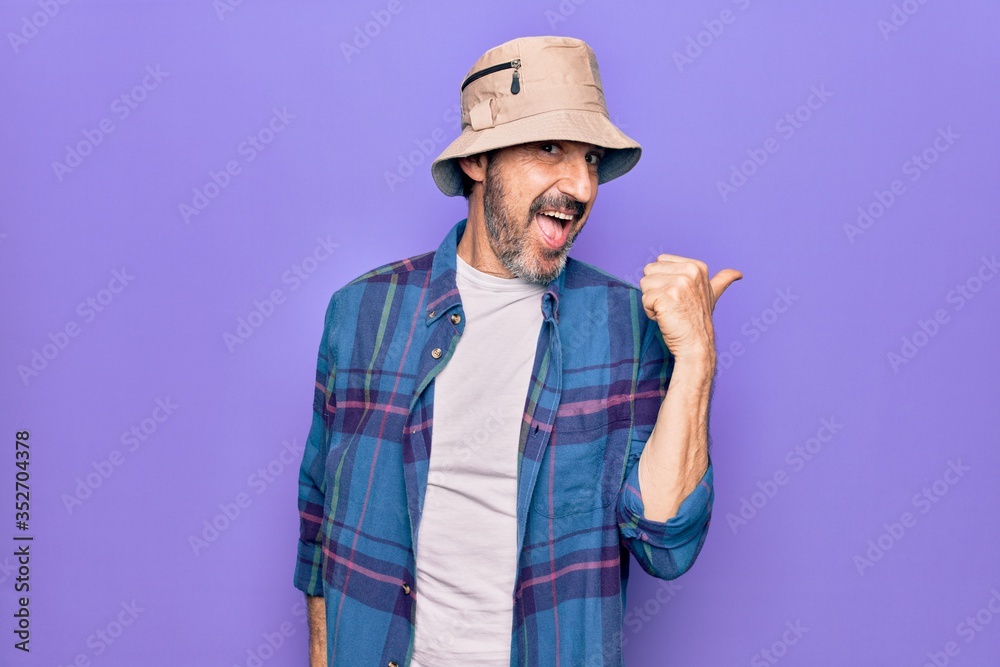 Middle age handsome man wearing casual shirt and hat over isolated purple background pointing thumb up to the side smiling happy with open mouth