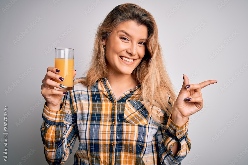 Young beautiful blonde woman drinking glass of healthy orange juice over white background very happy pointing with hand and finger to the side