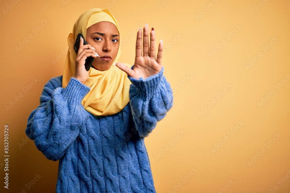 African American woman wearing muslim hijab having conversation talking on smartphone with open hand doing stop sign with serious and confident expression, defense gesture