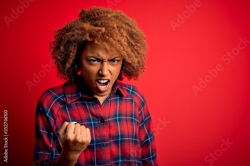 Young beautiful African American afro woman with curly hair wearing casual shirt angry and mad raising fist frustrated and furious while shouting with anger. Rage and aggressive concept. © Krakenimages.com