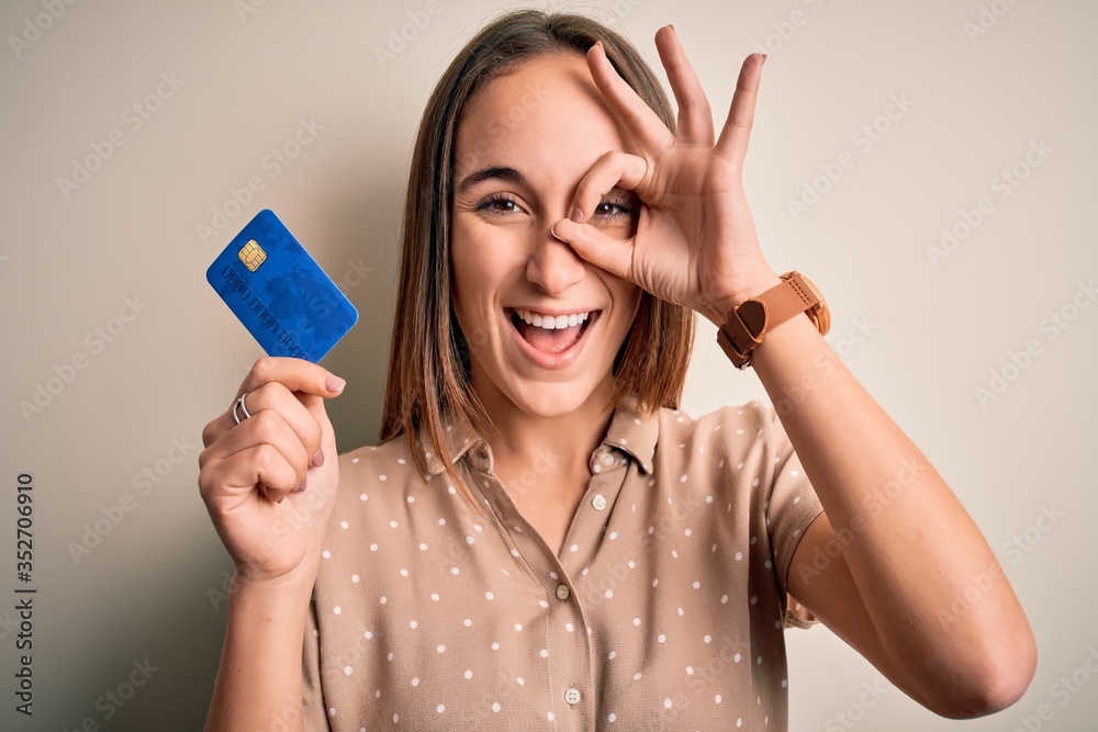 Young beautiful customer woman holding credit card to do payment over white background with happy face smiling doing ok sign with hand on eye looking through fingers Young beautiful customer woman holding credit card to do payment over white background with happy face smiling doing ok sign with hand on eye looking through fingers