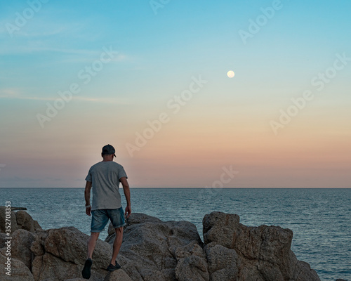 A young man in cap, t-shirt and jeans shirts walks on the rocks by the sea towards the moon