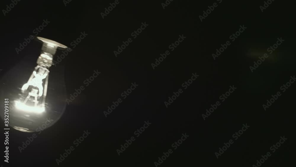 Classic glass lamp with tungsten filament lights up, swaying on the wire on a black background. Closeup. Slow mo, slo mo, slow motion, high speed camera