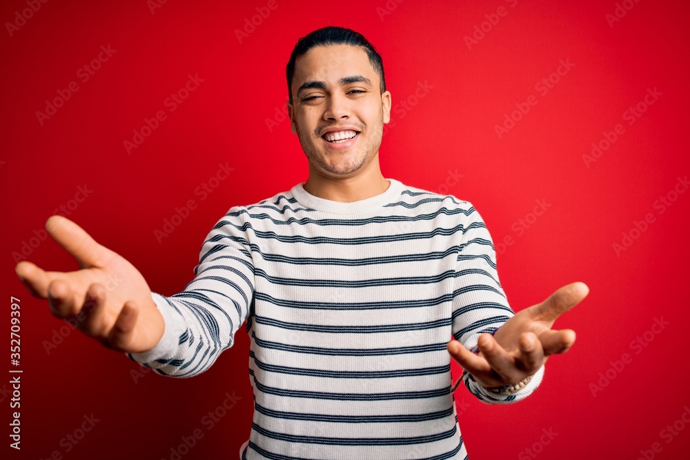 Fototapeta premium Young brazilian man wearing casual striped t-shirt standing over isolated red background smiling cheerful offering hands giving assistance and acceptance.
