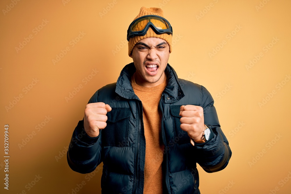 Young brazilian skier man wearing snow sportswear and ski goggles over yellow background angry and mad raising fists frustrated and furious while shouting with anger. Rage and aggressive concept. Young brazilian skier man wearing snow sportswear and ski goggles over yellow background angry and mad raising fists frustrated and furious while shouting with anger. Rage and aggressive concept.