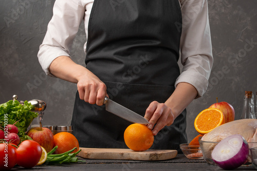 Chef cuts oranges for cooking dishes on the background of the ingredients of vegetables and fruits. Useful and proper nutrition