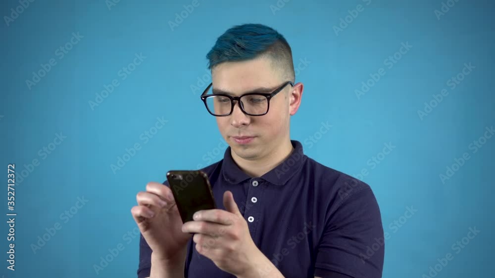 A young man in glasses with blue hair with a phone in his hands. An alternative person is writing in a smartphone on a blue background. A young man in glasses with blue hair with a phone in his hands. An alternative person is writing in a smartphone on a blue background.