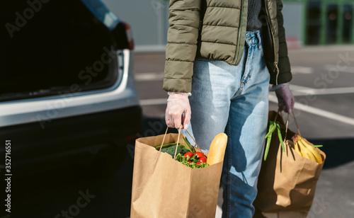A young woman with grocery bags from a supermarket. Car trunk at background. Social distancing: face mask, disposable gloves to prevent infection. Food shopping during coronavirus Covid 19 epidemic