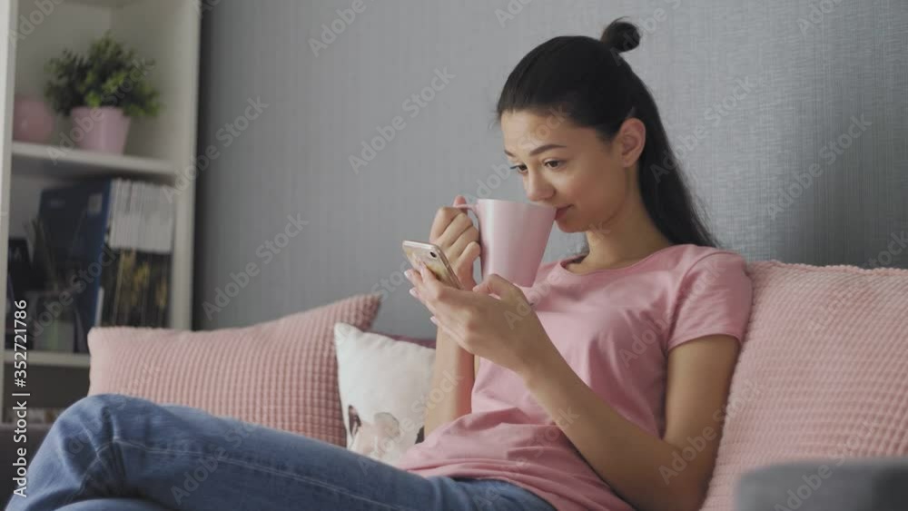 Beautiful young woman using smartphone relaxing on couch at home browsing online texting drinking coffee enjoying comfortable lifestyle Beautiful young woman using smartphone relaxing on couch at home browsing online texting drinking coffee enjoying comfortable lifestyle