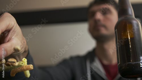 High quality close-up of a young blurred man chatting with friend or family on phone watching tv and having snacks with bottle of beer. Happy time. Handsome person with phone at home.