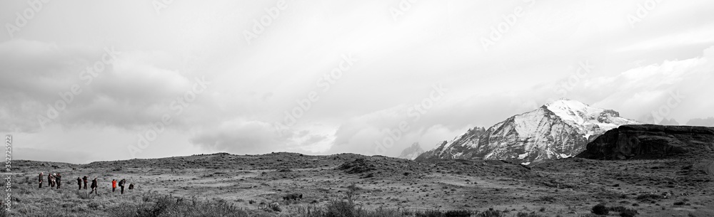 Black and white filter on Patagonia landscape.  Snow capped mountains on right panning to grassy meadow with herd of Guanaco, and line of Photographers.  Orange coat color splash. 