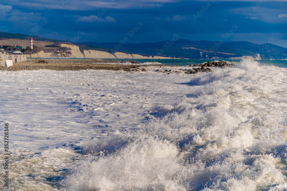In a storm, waves in snow-white foam fall on the pebble beach of Gelendzhik. In the background Gelendzhik lighthouse and rocks