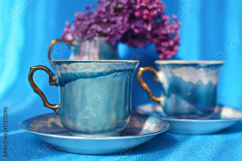 Beautiful couple blue porcelain cup of tea with golden handle on saucer on blurred background of cermic vase with blooming bunch of purple lilac. Selective focus. Horizontal frame