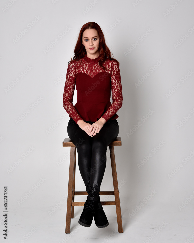 Portrait of a pretty girl with red hair wearing leather pants and long sleeved lace shirt.  full length sitting pose on wooden stool, isolated against a studio background