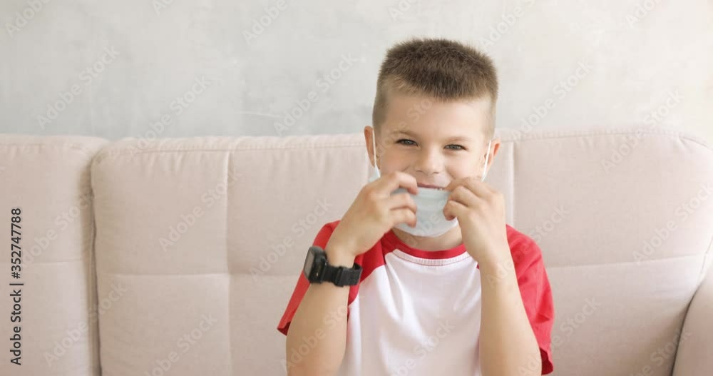 Portrait of smiling child boy wearing medical mask on quarantine sitting on sofa. He is playing with mask dressing it on face. Covid-19, coronavirus pandemic, epidemic, protective wear against virus. Portrait of smiling child boy wearing medical mask on quarantine sitting on sofa. He is playing with mask dressing it on face. Covid-19, coronavirus pandemic, epidemic, protective wear against virus.
