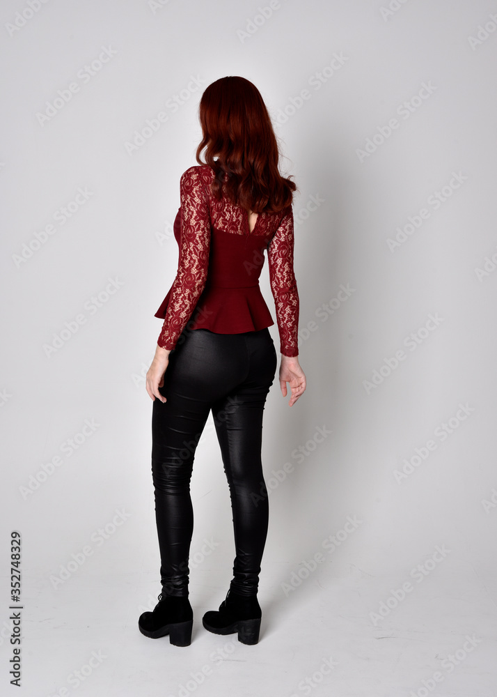 Portrait of a pretty girl with red hair wearing leather pants and long sleeved lace shirt. full length standing pose facing away from the camera, isolated against a studio background. Portrait of a pretty girl with red hair wearing leather pants and long sleeved lace shirt. full length standing pose facing away from the camera, isolated against a studio background.