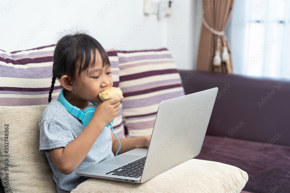 Asian little girl eating bread with using laptop, Studying homework online lesson at home Asian little girl eating bread with using laptop, Studying homework online lesson at home
