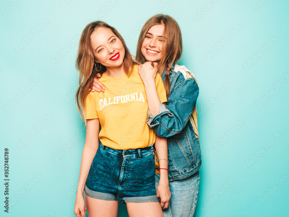 Two young beautiful smiling hipster girls in trendy summer yellow T-shirts and jeans jacket. Sexy carefree women posing near blue wall. Trendy and positive models having fun Two young beautiful smiling hipster girls in trendy summer yellow T-shirts and jeans jacket. Sexy carefree women posing near blue wall. Trendy and positive models having fun