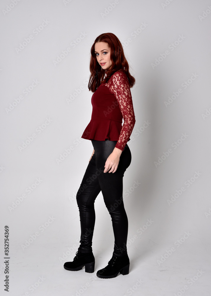 Portrait of a pretty girl with red hair wearing leather pants and long sleeved lace shirt. full length standing pose, isolated against a studio background. Portrait of a pretty girl with red hair wearing leather pants and long sleeved lace shirt. full length standing pose, isolated against a studio background.