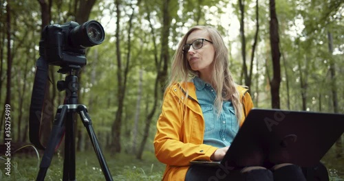 Wallpaper Mural Beautiful woman with blond hair sitting near tripod with digital camera and holding portable laptop on knees. Female photographer in yellow jacket and eyeglasses working with green nature around. Torontodigital.ca