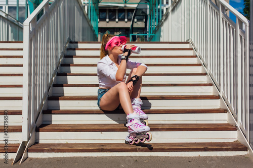 Portrait of an emotional girl in a pink cap visor wearing protective gloves and rollerblades sitting on stairs drinking water from a bottle