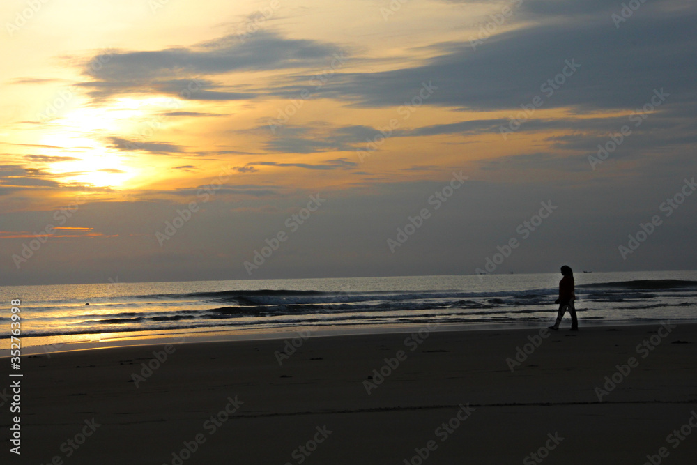 person / woman silhouette at the beach under yellow sunrise sun horizon. wavy beach. Dark foreground. tranquil lonely walk along the beach.