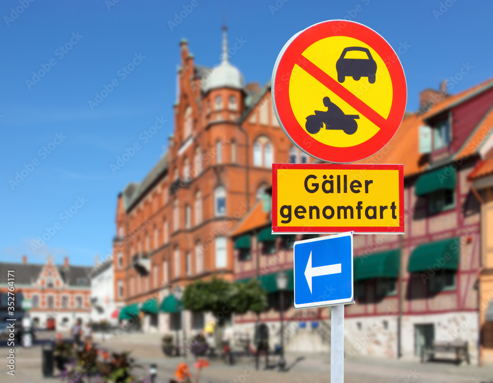 A yellow and red traffic sign in the Swedish port city of Ystad during the day in sunshine. The passage is prohibited for cars and motorbikes. Text translation: Applies to transit