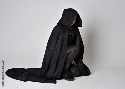 Fantasy portrait of a woman with red hair wearing dark leather assassin costume with long black cloak.  full length kneeling pose, isolated against a studio background.