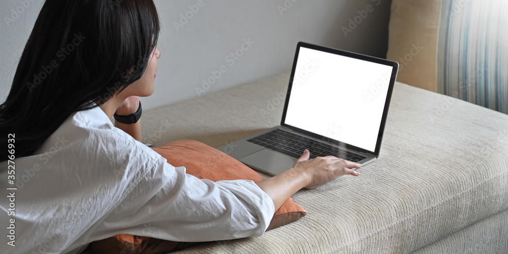 Photo of young woman laying on sofa and watching/using white blank screen computer laptop over comfortable living room as background. Woman with relaxing time concept. Photo of young woman laying on sofa and watching/using white blank screen computer laptop over comfortable living room as background. Woman with relaxing time concept.