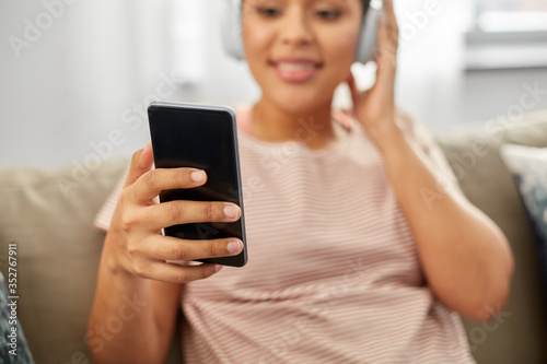 people, technology and leisure concept - close up of happy young african american woman in glasses with smartphone and headphones sitting on sofa and listening to music at home