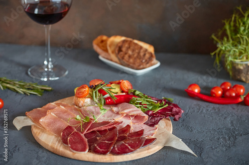 Assorted meat delicacies, cut into thin slices on a round wooden Board. Decorated with bruschetta with vegetables and fresh herbs