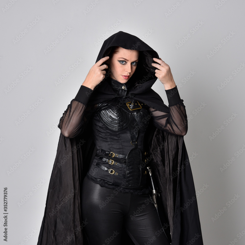 Close up fantasy portrait of a woman with red hair wearing dark leather assassin costume with long black cloak. isolated against a studio background. Close up fantasy portrait of a woman with red hair wearing dark leather assassin costume with long black cloak. isolated against a studio background.