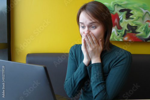 Girl with a perplexed surprised tense expression sits sitting working at a laptop at a table in a cafe on a brown sofa against a yellow wall. She put her hands on her face, covered her mouth with her